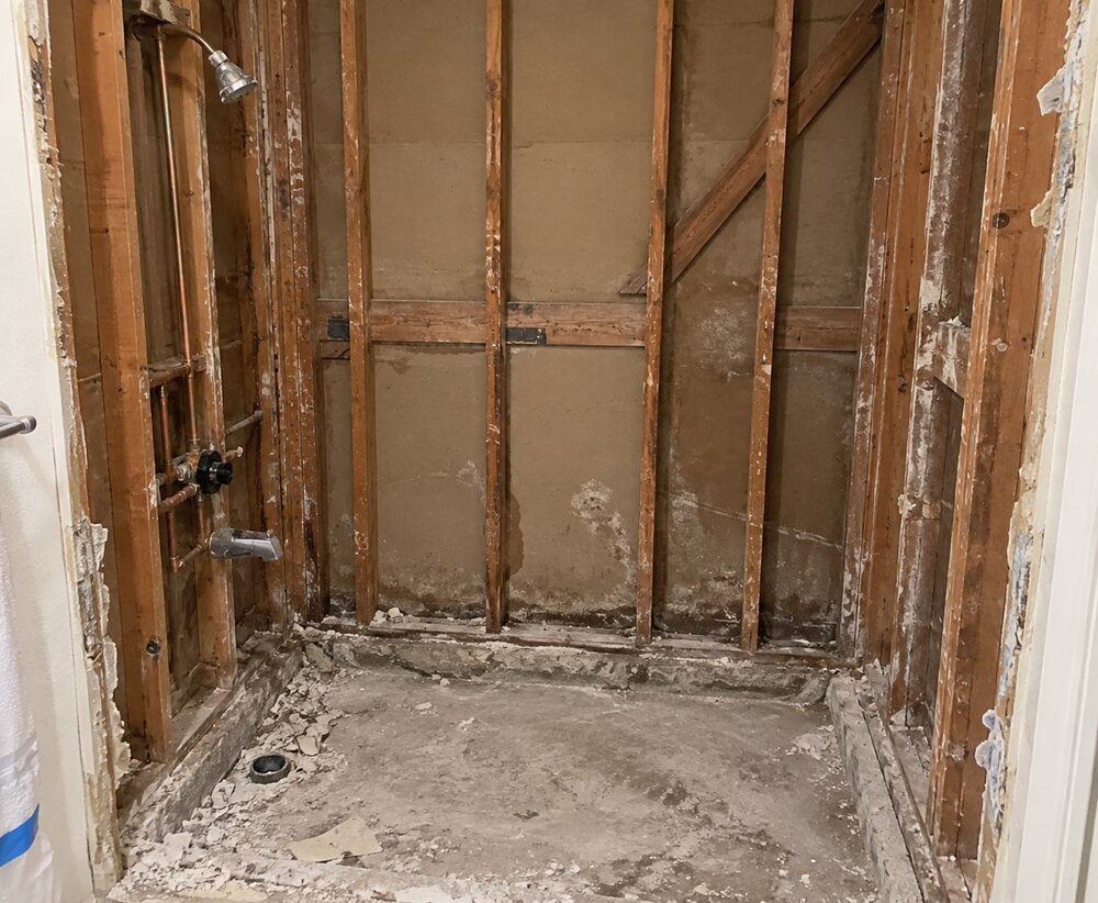 The same bathroom stripped to the studs, showing the old plumbing and the damaged framing before waterproofing